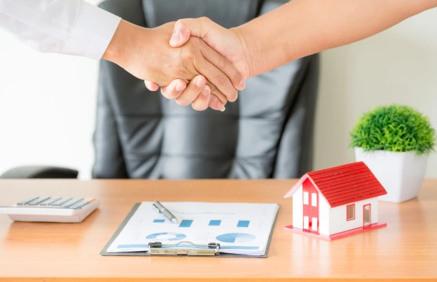 Handshake over desk with documents, house model, calculator, and a plant.